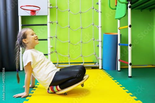 a little girl in a green room rides on a balance beam, treatment for disability in a children's rehabilitation center