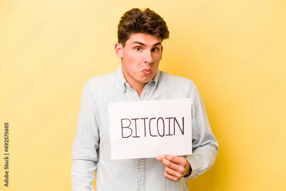 Young caucasian man holding bitcoin placard isolated on yellow background shrugs shoulders and open eyes confused. Young caucasian man holding bitcoin placard isolated on yellow background shrugs shoulders and open eyes confused.