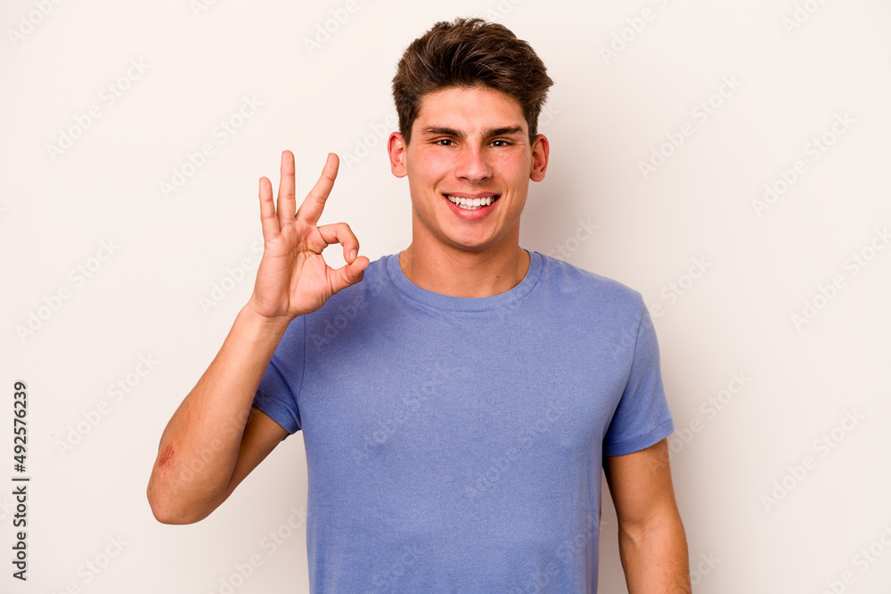 Young caucasian man isolated on white background winks an eye and holds an okay gesture with hand. Young caucasian man isolated on white background winks an eye and holds an okay gesture with hand.