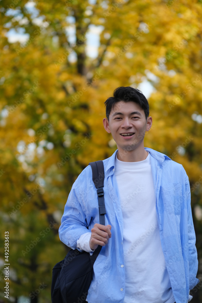 Asian male college student and blurred yellow ginkgo biloba tree in autumn. Carrying a shoulder bag and smiling at camera Asian male college student and blurred yellow ginkgo biloba tree in autumn. Carrying a shoulder bag and smiling at camera