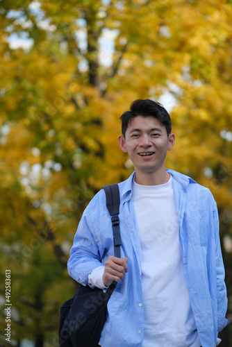 Asian male college student and blurred yellow ginkgo biloba tree in autumn. Carrying a shoulder bag and smiling at camera