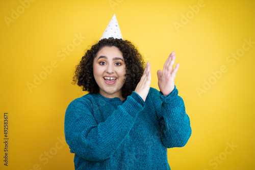 Young beautiful woman wearing a birthday hat over isolated yellow background clapping and applauding happy and joyful, smiling proud hands together © Irene