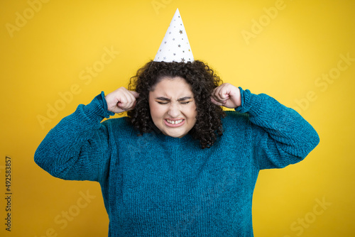 Young beautiful woman wearing a birthday hat over isolated yellow background covering ears with fingers with annoyed expression for the noise of loud music. Deaf concept. © Irene