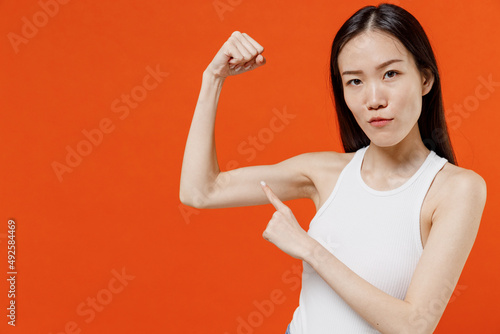 Confident young woman of Asian ethnicity 20s years old in white tank top showing pointing forefinger on biceps muscles demonstrating strength power isolated on plain orange background studio portrait.