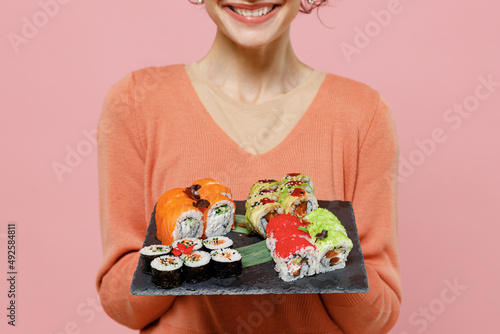 Cropped young smiling happy fun cool woman 20s wearing casual clothes holding in hand makizushi sushi roll served on black plate traditional japanese food isolated on plain pastel pink background