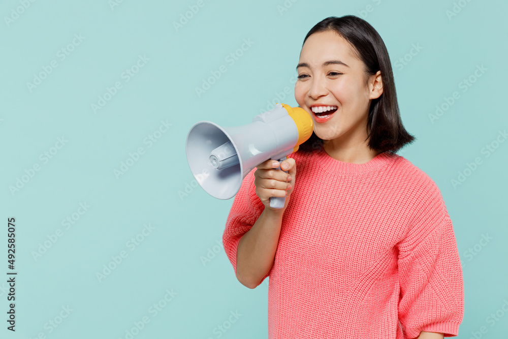 Young smiling happy woman of Asian ethnicity 20s wearing pink sweater hold scream in megaphone announces discounts sale Hurry up isolated on pastel plain light blue color background studio portrait.