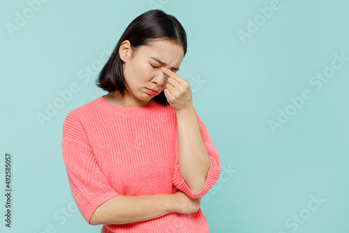 Young pensive sad woman of Asian ethnicity 20s wearing pink sweater keep eyes closed rub put hand on nose isolated on pastel plain light blue color background studio portrait People lifestyle concept
