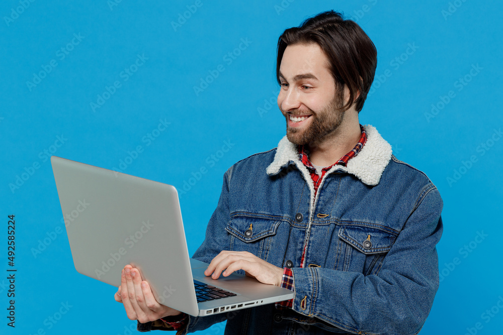 Smiling happy cheery young brunet bearded man 20s wear warm denim jacket hold use work on laptop pc computer isolated on plain pastel light blue background studio portrait. Winter cold season concept Smiling happy cheery young brunet bearded man 20s wear warm denim jacket hold use work on laptop pc computer isolated on plain pastel light blue background studio portrait. Winter cold season concept