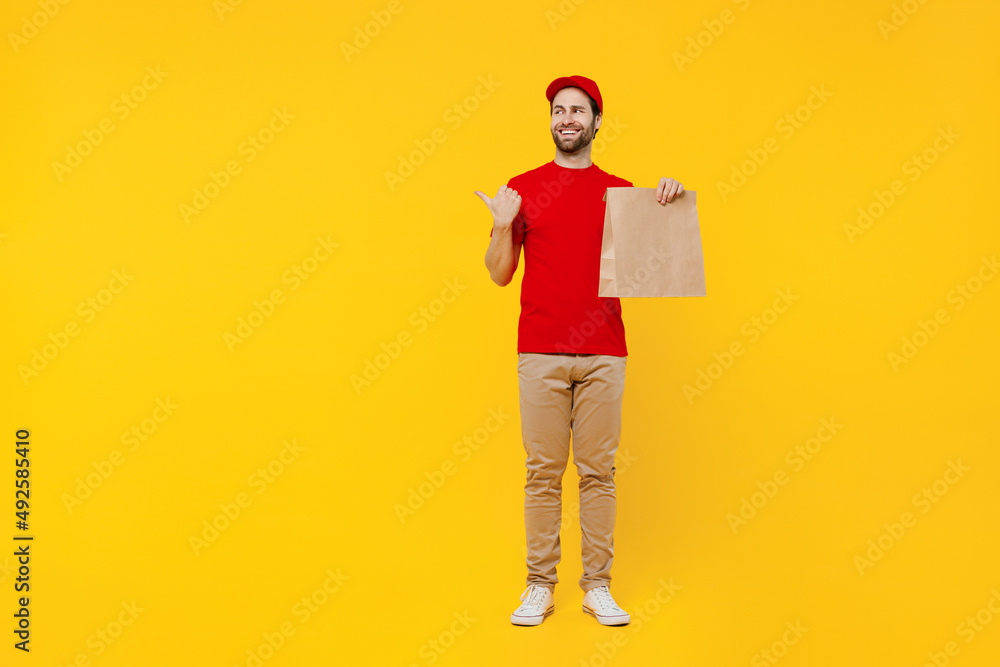 Full body delivery guy employee man in red cap T-shirt uniform workwear work as dealer courier hold brown blank craft paper takeaway bag mock up point finger aside isolated on plain yellow background. Full body delivery guy employee man in red cap T-shirt uniform workwear work as dealer courier hold brown blank craft paper takeaway bag mock up point finger aside isolated on plain yellow background.