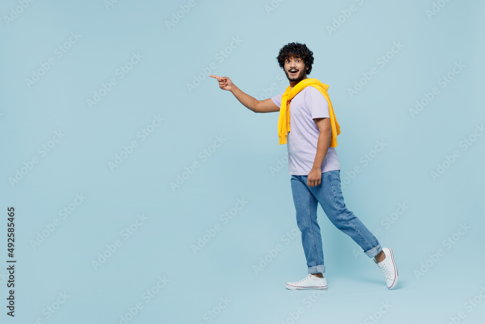 Full size amazed fancy young bearded Indian man 20s wears white t-shirt pointing index finger aside on workspace area copy space mock up isolated on plain pastel light blue background studio portrait.
