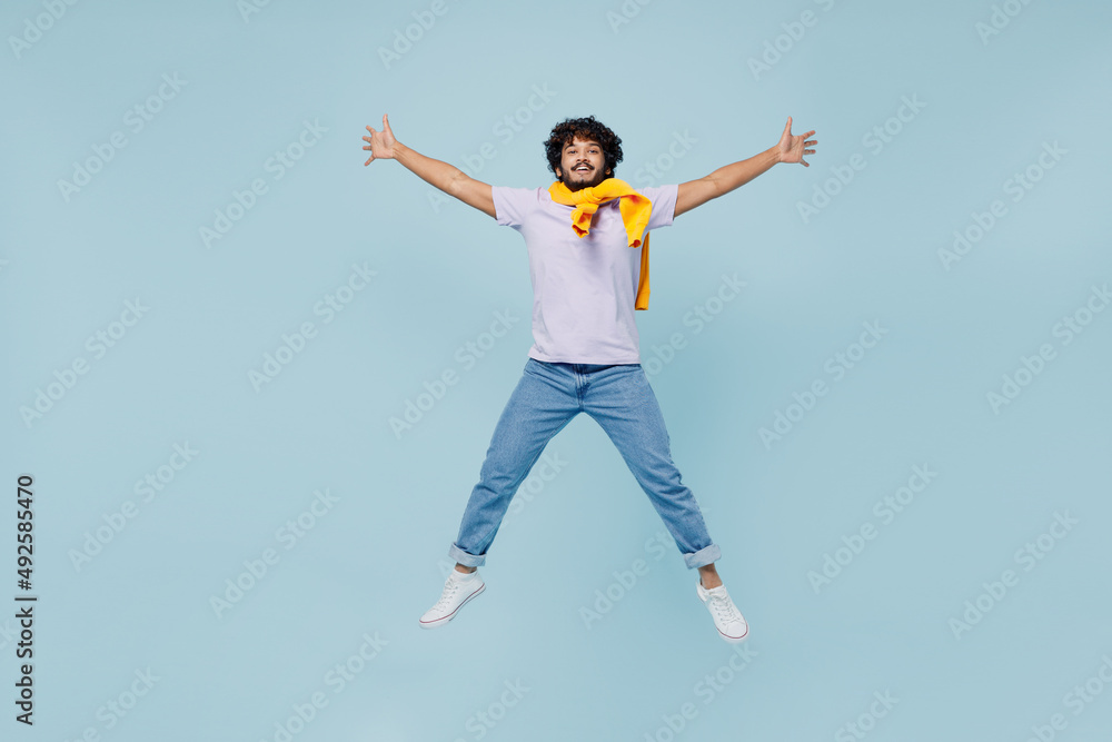 Full size body length cheerful happy young bearded Indian man 20s years old wears white t-shirt raise up spreading hands looking camera isolated on plain pastel light blue background studio portrait. Full size body length cheerful happy young bearded Indian man 20s years old wears white t-shirt raise up spreading hands looking camera isolated on plain pastel light blue background studio portrait.