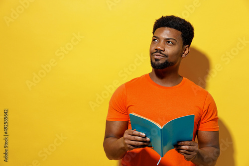Young minded fun student man of African American ethnicity 20s wear orange t-shirt read novel book learn look aside dream isolated on plain yellow background studio portrait. People lifestyle concept.