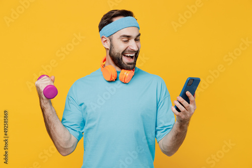 Young smiling happy fitness trainer instructor sporty man sportsman wear headband blue t-shirt hold dumbbells mobile cell phone isolated on plain yellow background. Workout sport motivation concept