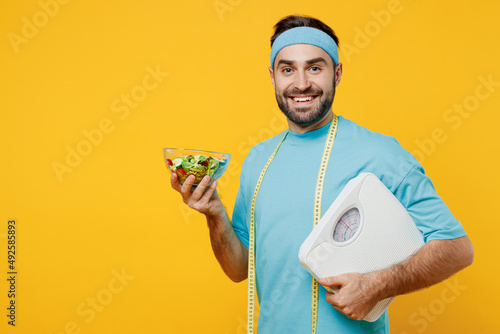 Young smiling fitness trainer instructor sporty man sportsman in headband blue t-shirt measure tape hold plate with vitamin salad hug scales isolated on plain yellow background. Workout sport concept. photo