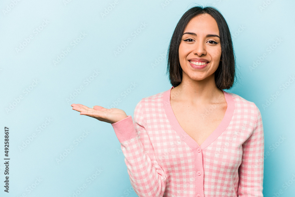 Young hispanic woman isolated on blue background showing a copy space on a palm and holding another hand on waist. Young hispanic woman isolated on blue background showing a copy space on a palm and holding another hand on waist.