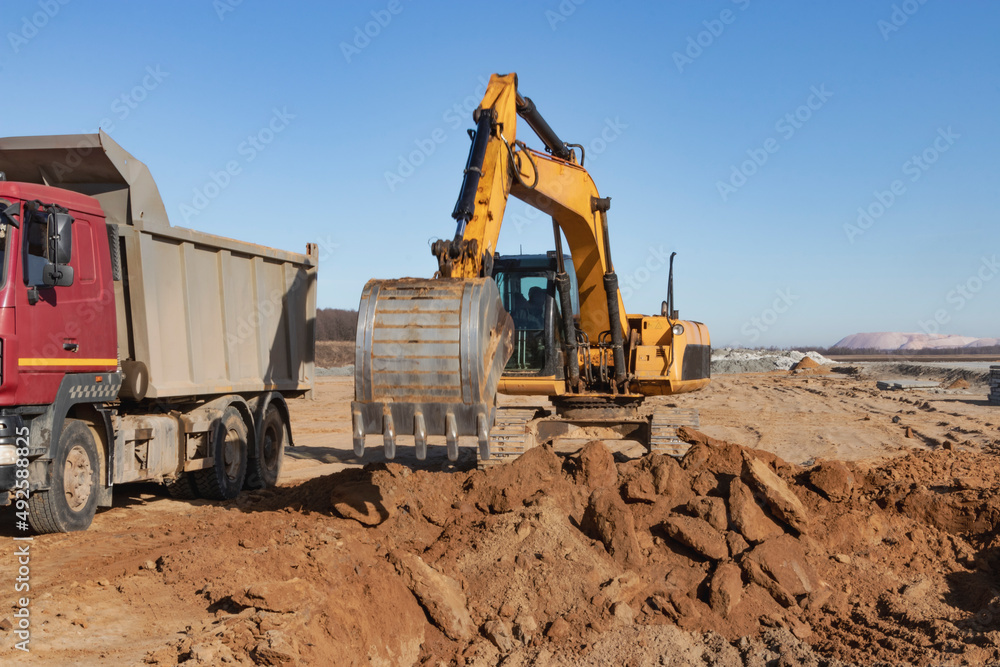 A powerful crawler excavator loads the earth into a dump truck against the blue sky. Development and removal of soil from the construction site. A powerful crawler excavator loads the earth into a dump truck against the blue sky. Development and removal of soil from the construction site.