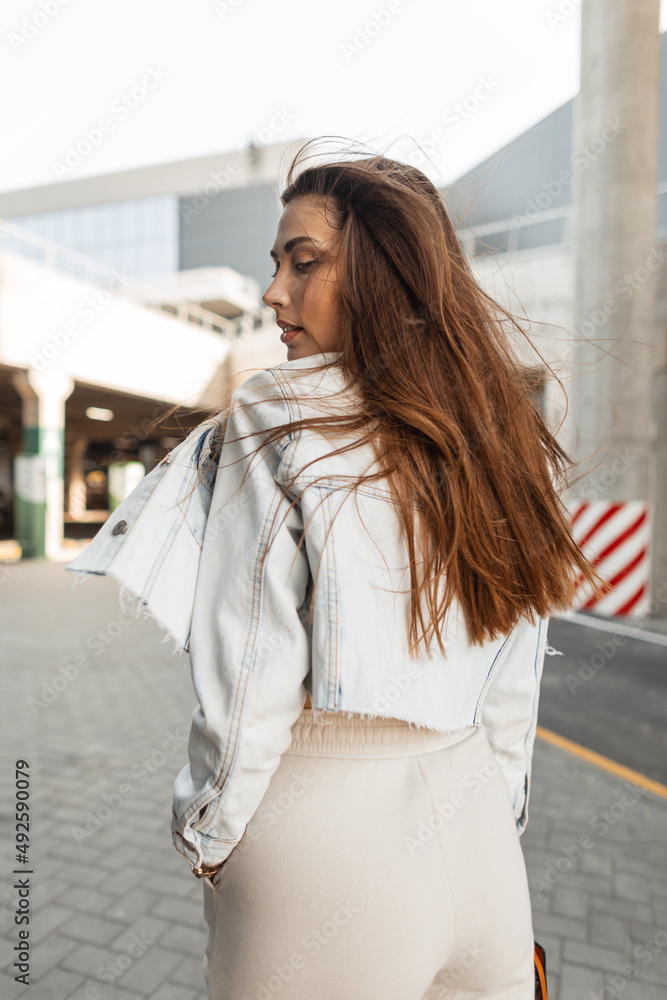 Young beautiful hipster girl with brunette hair in a fashionable denim jacket walks in the city near a modern building Young beautiful hipster girl with brunette hair in a fashionable denim jacket walks in the city near a modern building