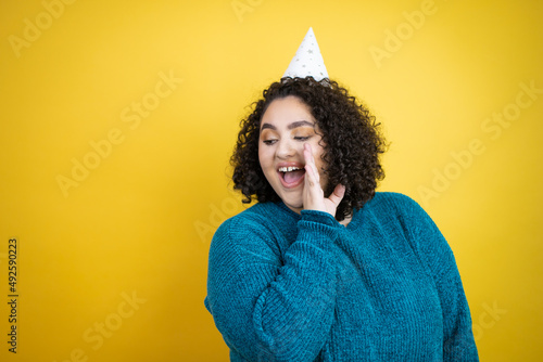 Young beautiful woman wearing a birthday hat over isolated yellow background hand on mouth telling secret rumor, whispering malicious talk conversation © Irene