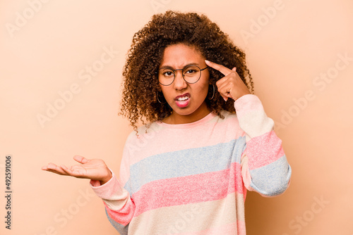 Young African American woman isolated on beige background holding and showing a product on hand.