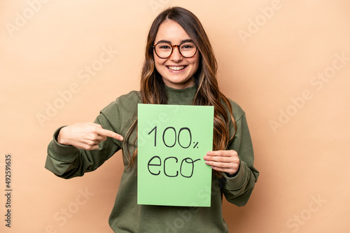 Young caucasian woman holding 100% eco placard isolated on beige background person pointing by hand to a shirt copy space, proud and confident