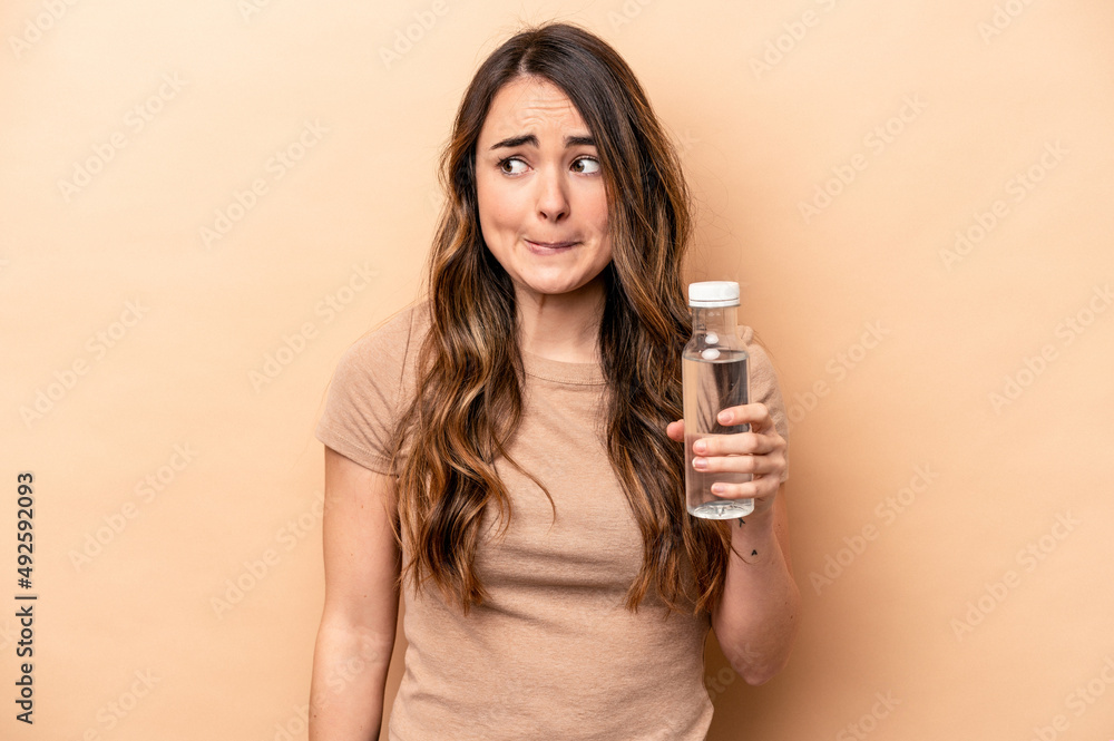Young caucasian woman holding a bottle of water isolated on beige background confused, feels doubtful and unsure. Young caucasian woman holding a bottle of water isolated on beige background confused, feels doubtful and unsure.