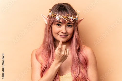 Young elf woman with pink hair isolated on beige background pointing with finger at you as if inviting come closer.