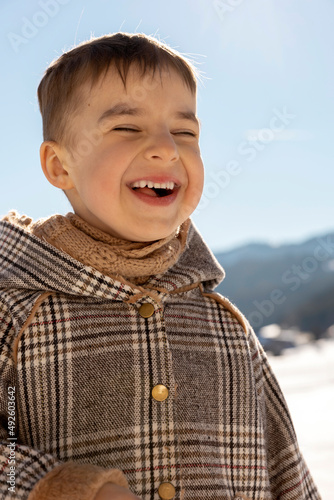 Portrait of little adorable boy with winter landscape in the background. Cute, smiling caucasian child with brown coat standing outdoor and enjoying good weather, blue sky, snow. Good, positive mood.