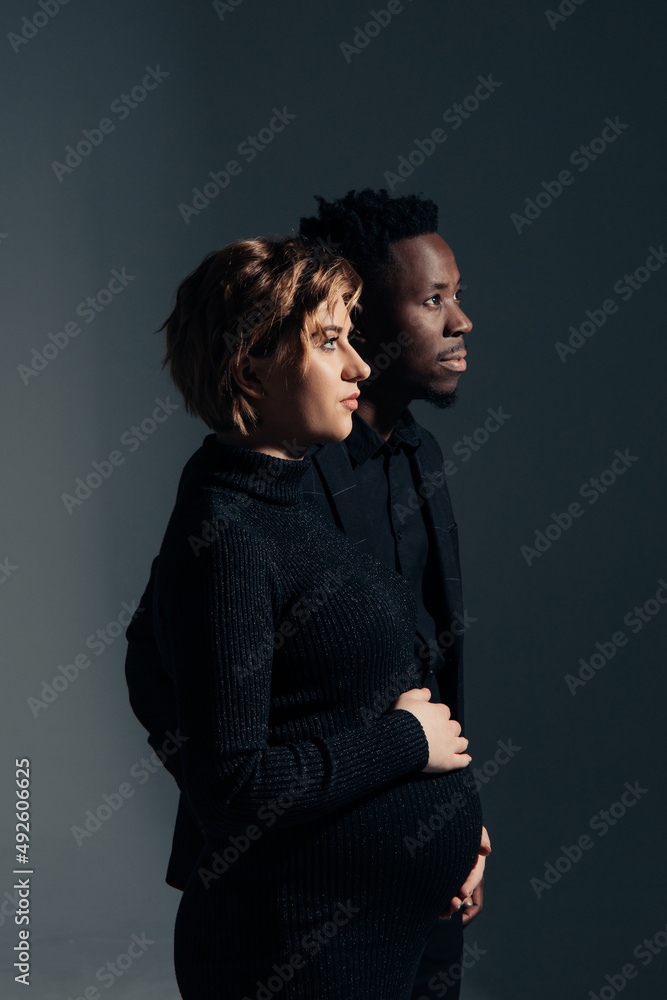 Young african man and his caucasian pregnant woman stand against grey background.