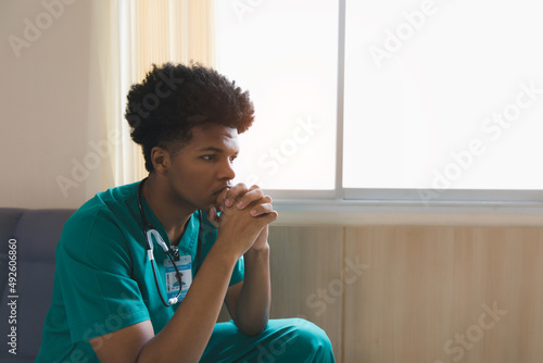 Shot of a young doctor looking distressed. Tired exhausted male african scrub nurse wears green uniform gloves sits on hospital. Depressed sad black ethic doctor feels burnout stress