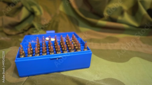 Ammunition box full of powerful military grade 223 caliber varmint bullets with blue plastic tip laying on camouflage jacket - Slider from left to right