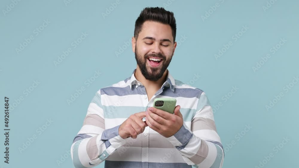 Boring uninspired dull tedious young bearded brunet man 20s years old wears striped shirt take mobile cell phone use laugh read search isolated on plain pastel light blue background studio portrait Boring uninspired dull tedious young bearded brunet man 20s years old wears striped shirt take mobile cell phone use laugh read search isolated on plain pastel light blue background studio portrait