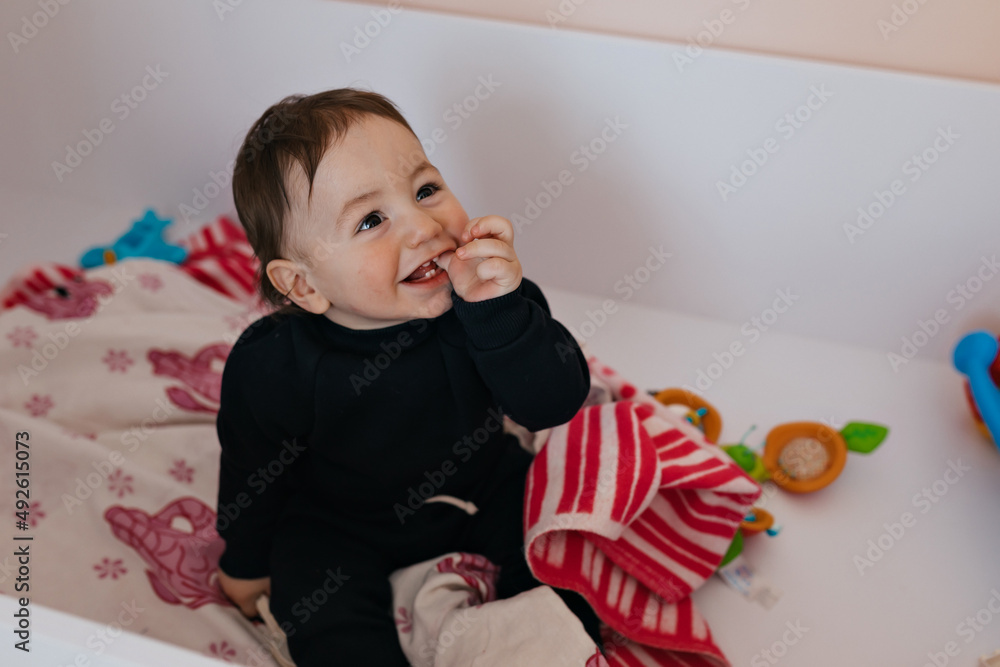 Lovely happy little boy in home clothes is sitting in his bed and looking up with happy smile. Child playing with colorful toy blocks. Kids play. Little boy in sunny white bedroom. Lovely happy little boy in home clothes is sitting in his bed and looking up with happy smile. Child playing with colorful toy blocks. Kids play. Little boy in sunny white bedroom.