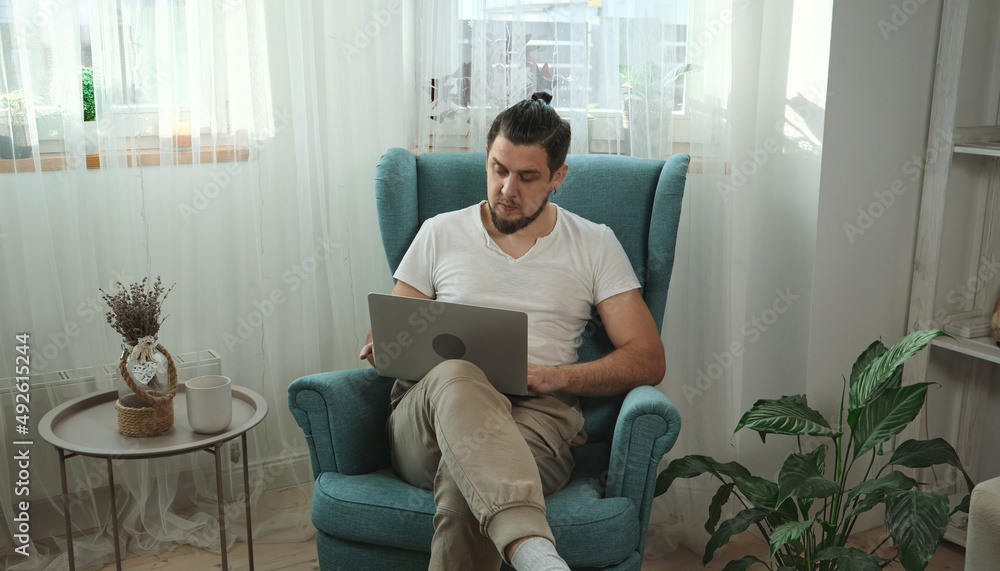 Young man using laptop typing and working sitting on the armchair at home