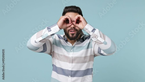 Smiling happy funny young bearded brunet man 20s years old wears striped shirt holding hands near eyes imitating glasses or binoculars isolated on plain pastel light blue background studio portrait