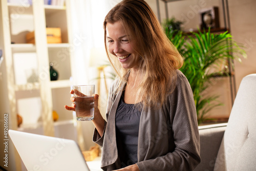 Girl holding a glass of water. Smiling girl drinking water while using the laptop..