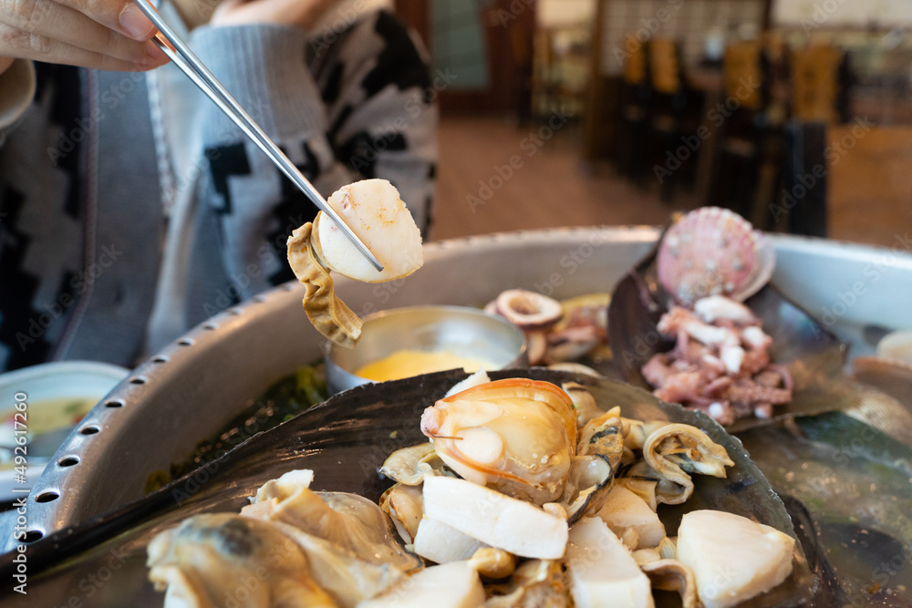 A woman is holding a slice of boiled clams among seafood served in a large clams shell.