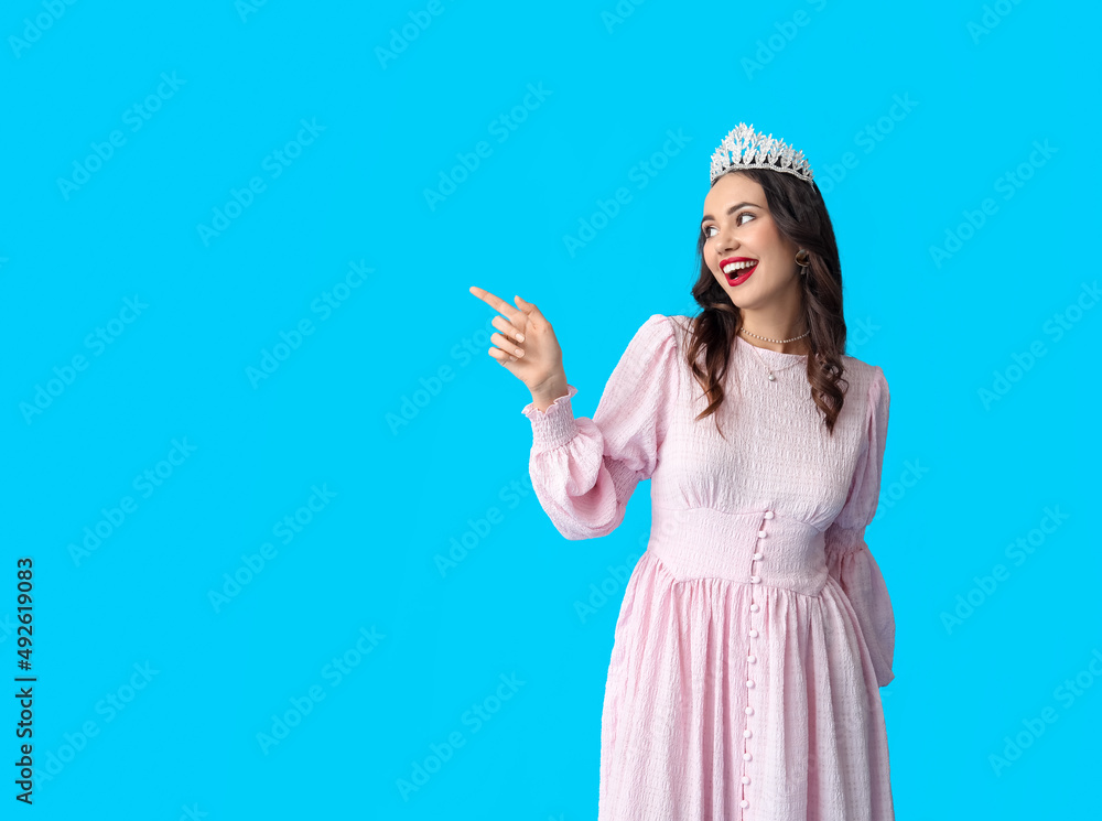 Beautiful young woman in stylish dress and tiara pointing at something on blue background Beautiful young woman in stylish dress and tiara pointing at something on blue background