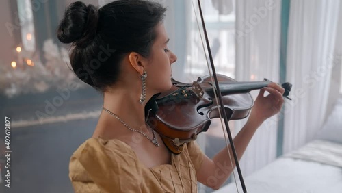 A Young Girl And Man Plays The Violin In A Beautiful Retro Room With Flowers And A Piano.