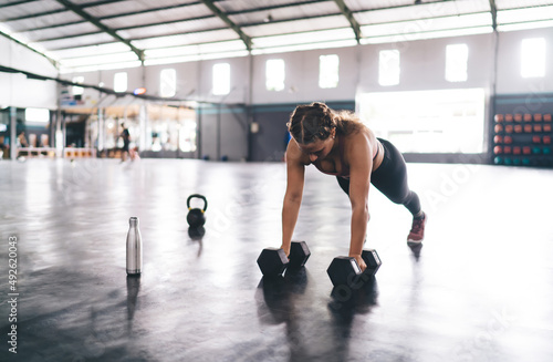Strong woman in sportive wear doing push up exercises during morning workout in gym studio, determined female trainer in tracksuit practice plank with dumbbells keeping body shape with physical effort