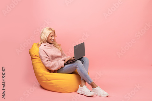 Freelance career. Happy young woman working with laptop computer, sitting in beanbag chair, pink background, copy space photo