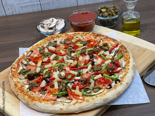 Not cut veggie pizza on wood board on table and pizza cutter and fresh mushrooms and olive oil on side.