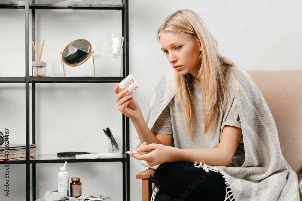 The girl is looking at the medicine and deciding what to take. Young woman has sore throat and high temperature. Concept of health