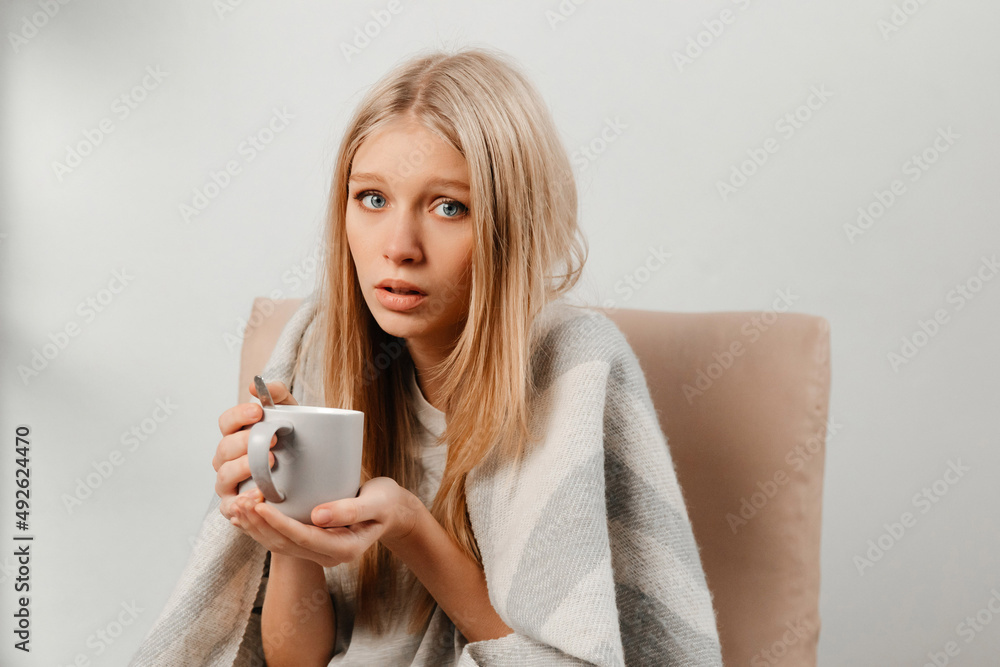 Nice-looking girl is looking at the camera and holding a cup of hot herbal tea. The girl is cold and trying to get warmer. Young woman is ill and taking care of her health. Concept of health Nice-looking girl is looking at the camera and holding a cup of hot herbal tea. The girl is cold and trying to get warmer. Young woman is ill and taking care of her health. Concept of health