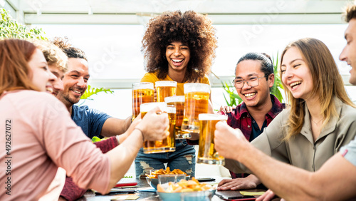 Mixed race group of friends toasting beer glasses at brewery pub - Happy young people having indoor dinner party celebrating summertime together - Beverage, food and friendship lifestyle concept