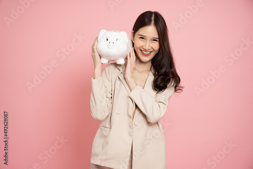 Portrait of Happy Asian businesswoman holding white piggy bank isolated on pink background, Saving money and financial economize concept photo