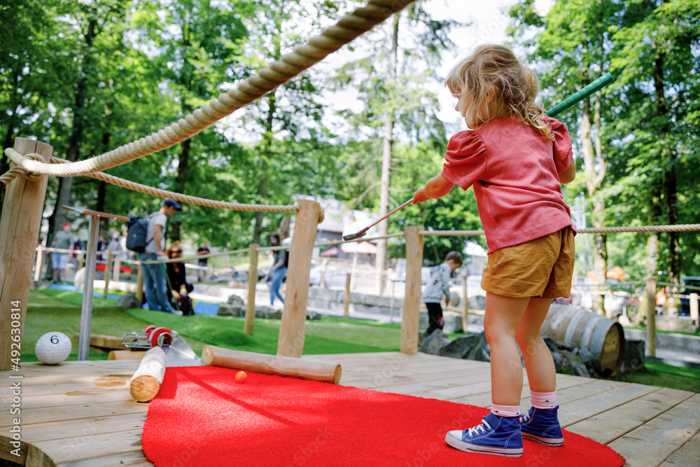 Cute preschool girl playing mini golf with family. Happy toddler child having fun with outdoor activity. Summer sport for children and adults, outdoors. Family vacations or resort. - obrazy, fototapety, plakaty 