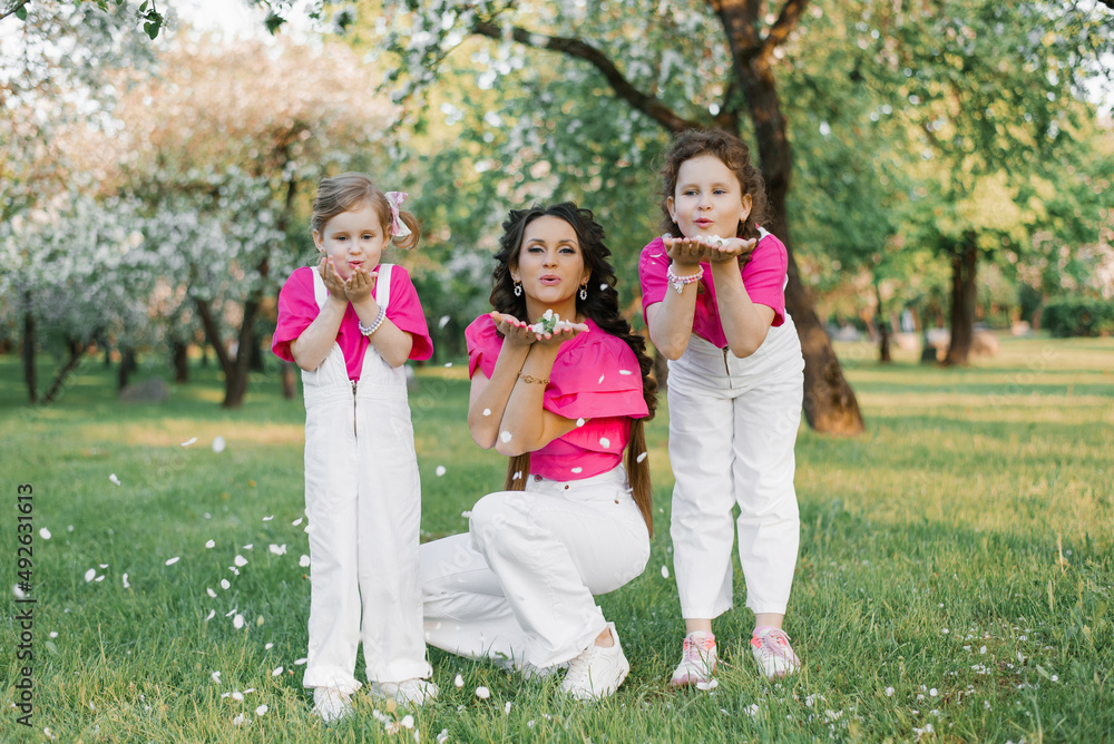 Obraz premium Cute mom and two daughters blowing on the fallen petals of apple blossoms in the spring garden. Outdoor activity. Sweet moments family