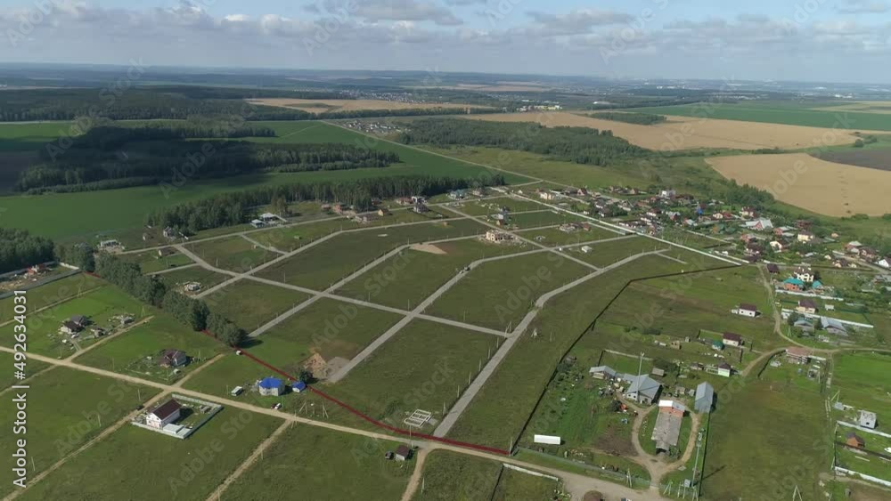 Aerial view of vacant land plots in new cottage village. There are dirt roads in the village, houses and electric poles are being built. Around the meadows, fields and forest. There are clouds in the Aerial view of vacant land plots in new cottage village. There are dirt roads in the village, houses and electric poles are being built. Around the meadows, fields and forest. There are clouds in the