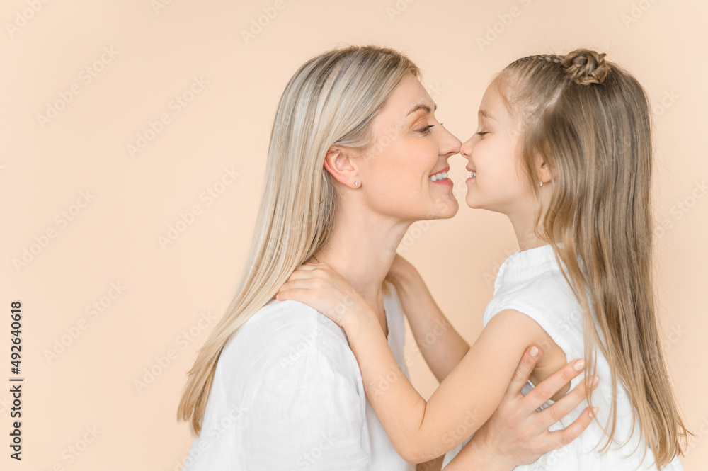 Mom and little daughter on a beige background in white clothes. Nose to nose. Mom and little daughter on a beige background in white clothes. Nose to nose.