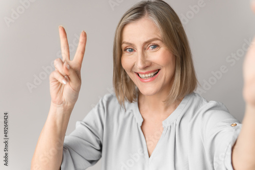 Charmind age lady waves to the interlocutor on video talk. Close up self-portrait picture of smiling old woman on light background. photo